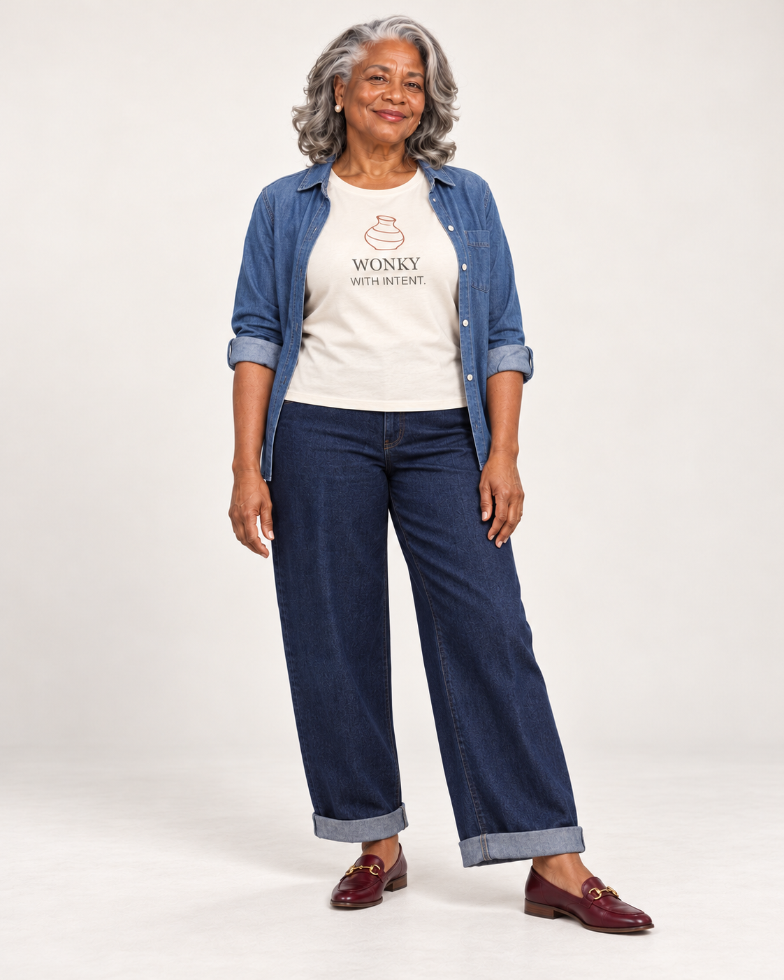 Vivienne wearing the Natural Wonky With Intent Bella+Canvas 6400 women’s relaxed-fit tee with an open blue overshirt, dark cuffed jeans and burgundy loafers.
